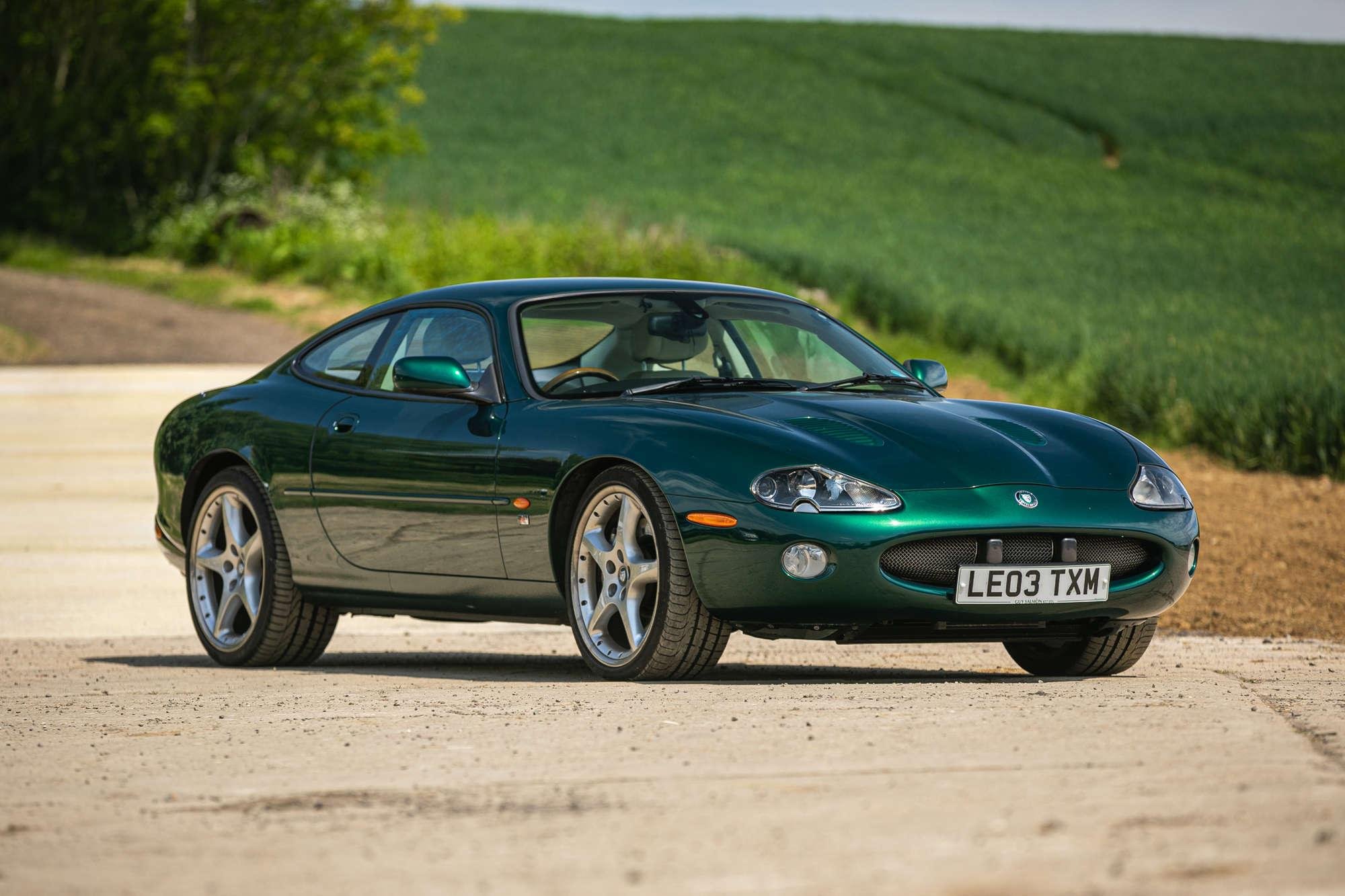 A green sports car parked on a gravel road with green fields and a tree in the background.