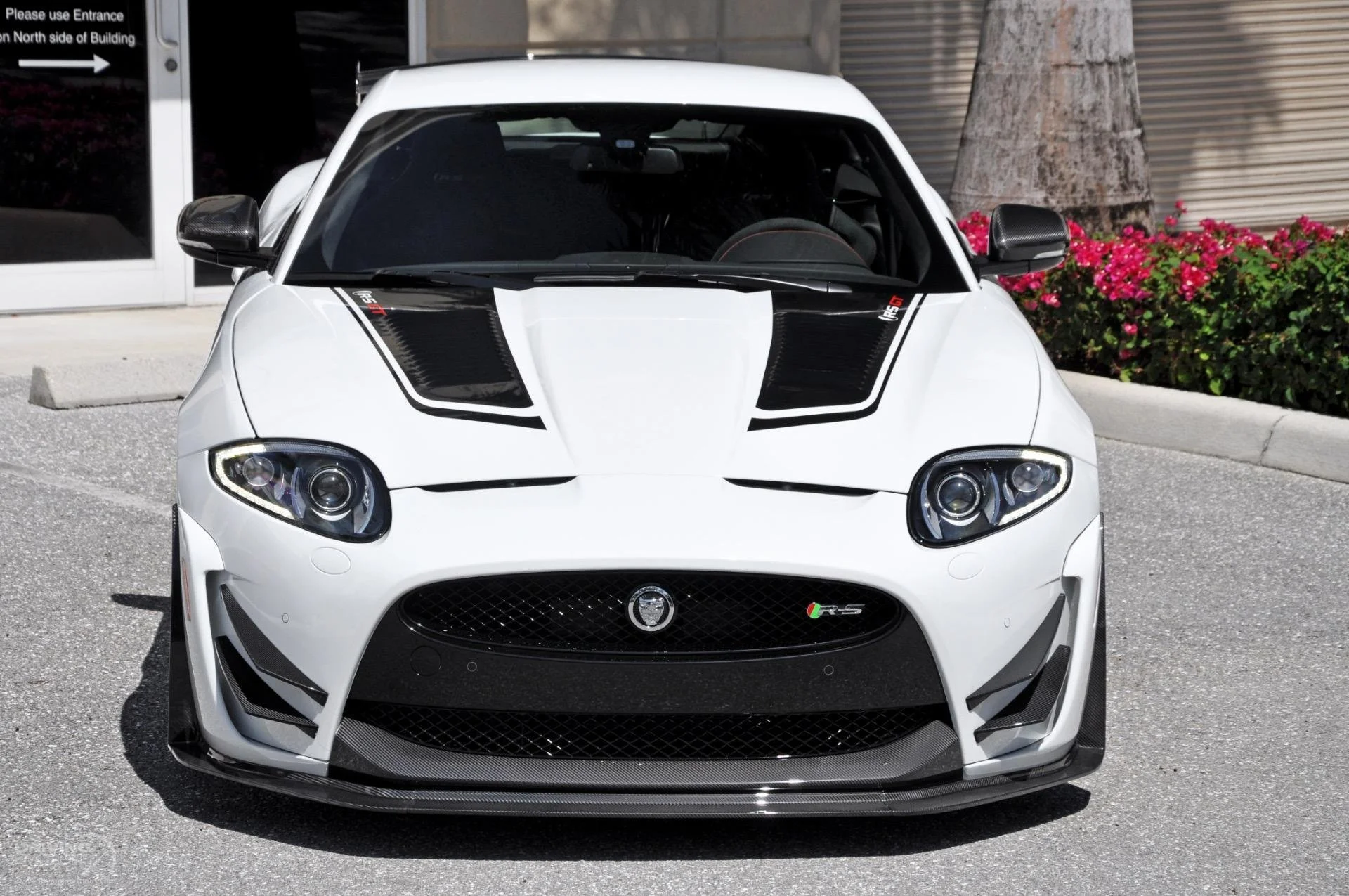 Front view of a white Jaguar F-Type sports car parked outdoors, with a black racing stripe on the hood and features the Jaguar emblem on the grille.