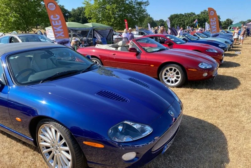 A line of classic sports cars, including a blue and red convertible, displayed outdoors at a car show with tents and people in the background.