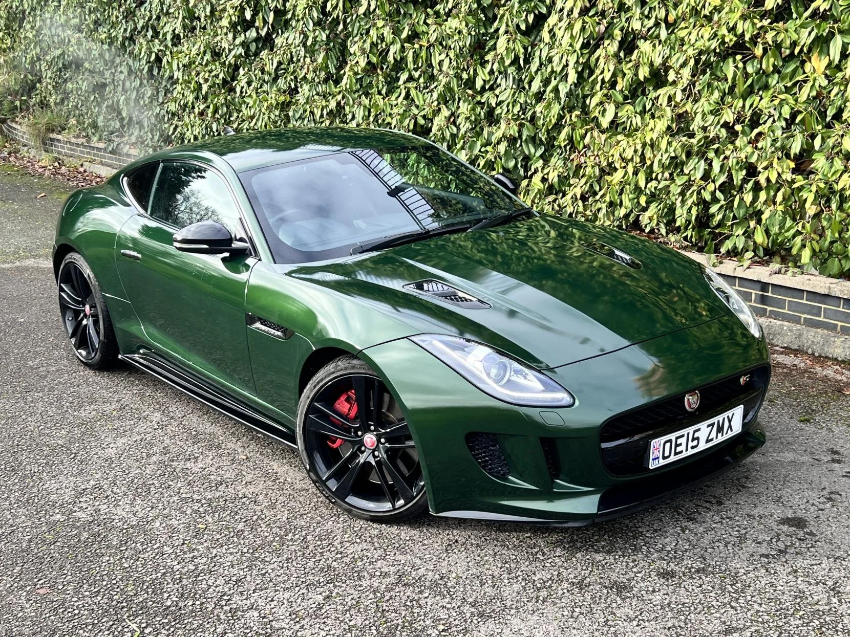 A green sports car, likely a Jaguar F-Type, parked on a paved surface beside a garden hedge. The car has black wheels with red brake calipers, and a UK license plate reading 'OE15 ZMX'.