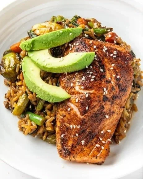 Cooked salmon fillet served on a bed of rice and vegetables, topped with sliced avocado and sprinkled with salt.