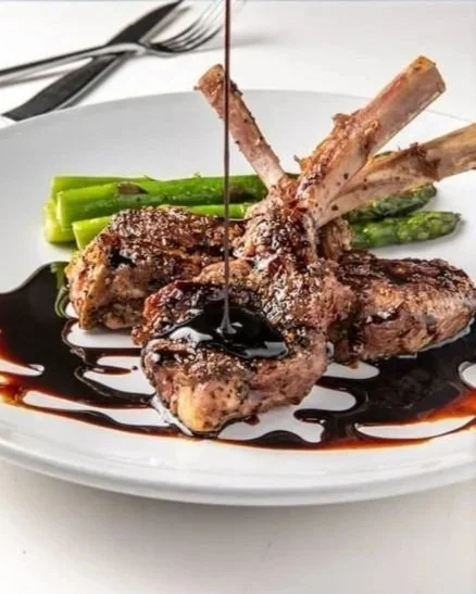 A plate of cooked lamb chops garnished with green asparagus, drizzled with a dark sauce, with some utensils in the background.