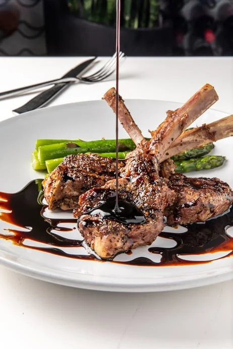 A plate of grilled lamb chops with asparagus and balsamic glaze, with a fork and knife on a white table.