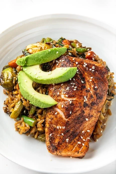 Grilled salmon fillet with sesame seeds served on a bed of mixed vegetables and brown rice, garnished with sliced avocado and lime wedges.