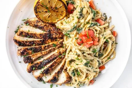 Grilled chicken breast with seasoned spices, mashed potatoes with herbs, spaghetti pasta with tomato chunks and herbs, and a grilled lemon half