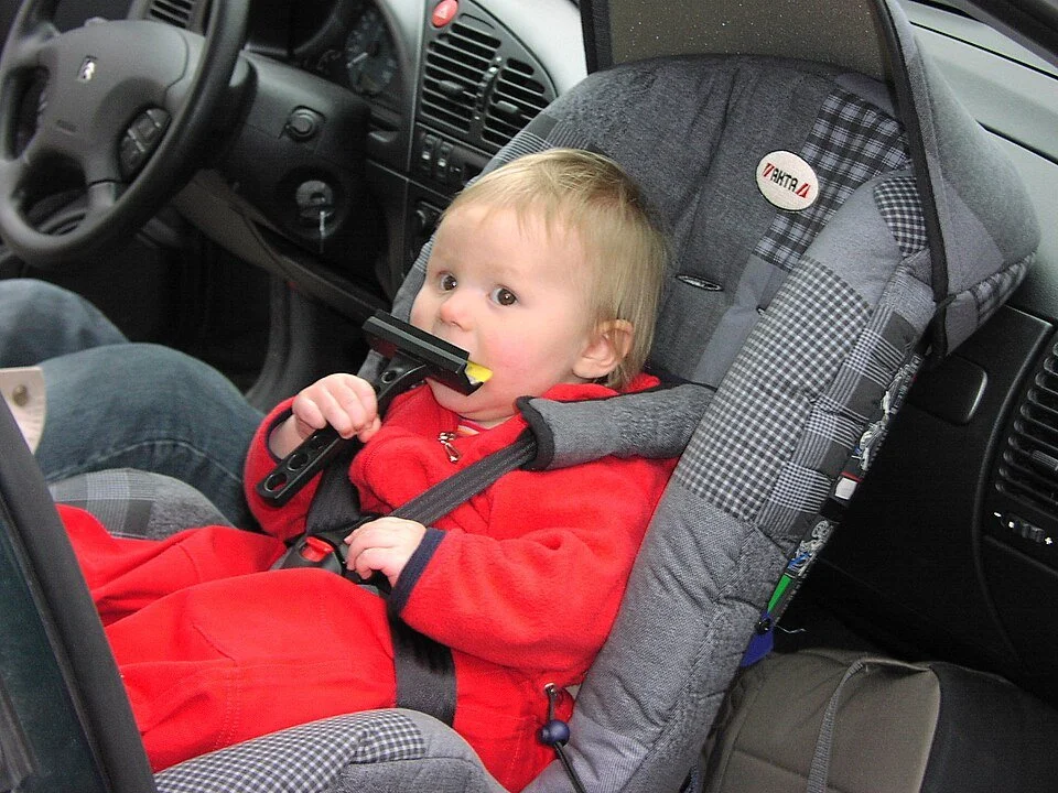 A young child with blond hair sitting in a car seat in the front passenger seat of a vehicle, holding a toy gun to their mouth.
