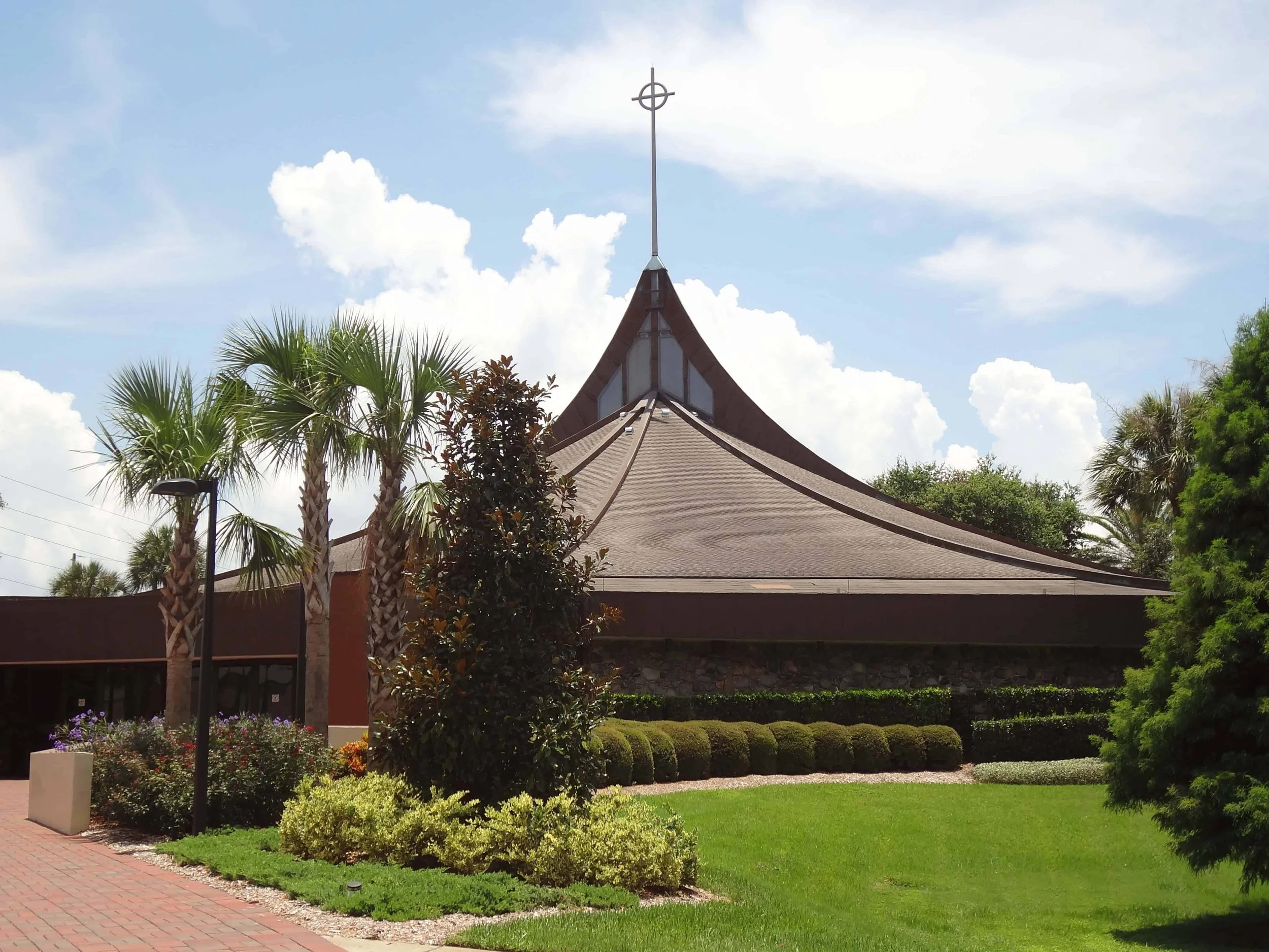 A church with a tall steeple topped by a cross, surrounded by palm trees, bushes, and a grass lawn, under a sky with clouds.