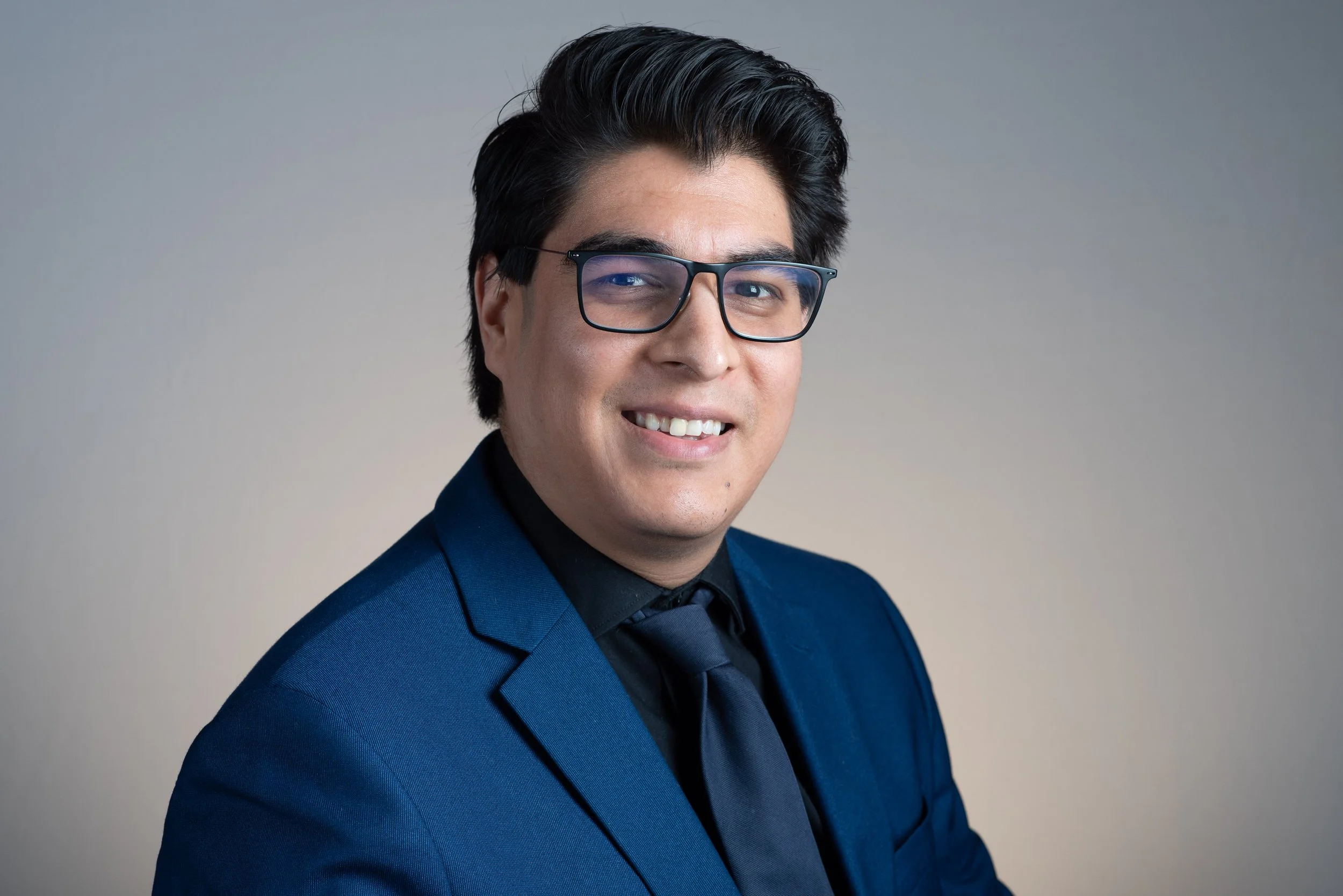 A young man with black hair slicked back, wearing black-rimmed glasses and a blue suit, smiling and looking at the camera, against a plain gray background.