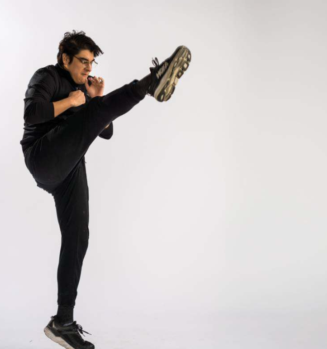 Man performing a high kick in a martial arts pose in a studio setting with a plain white background.