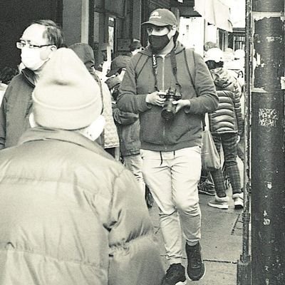 Person wearing a mask, hat, and hoodie holding a camera on a busy city street with pedestrians and buildings