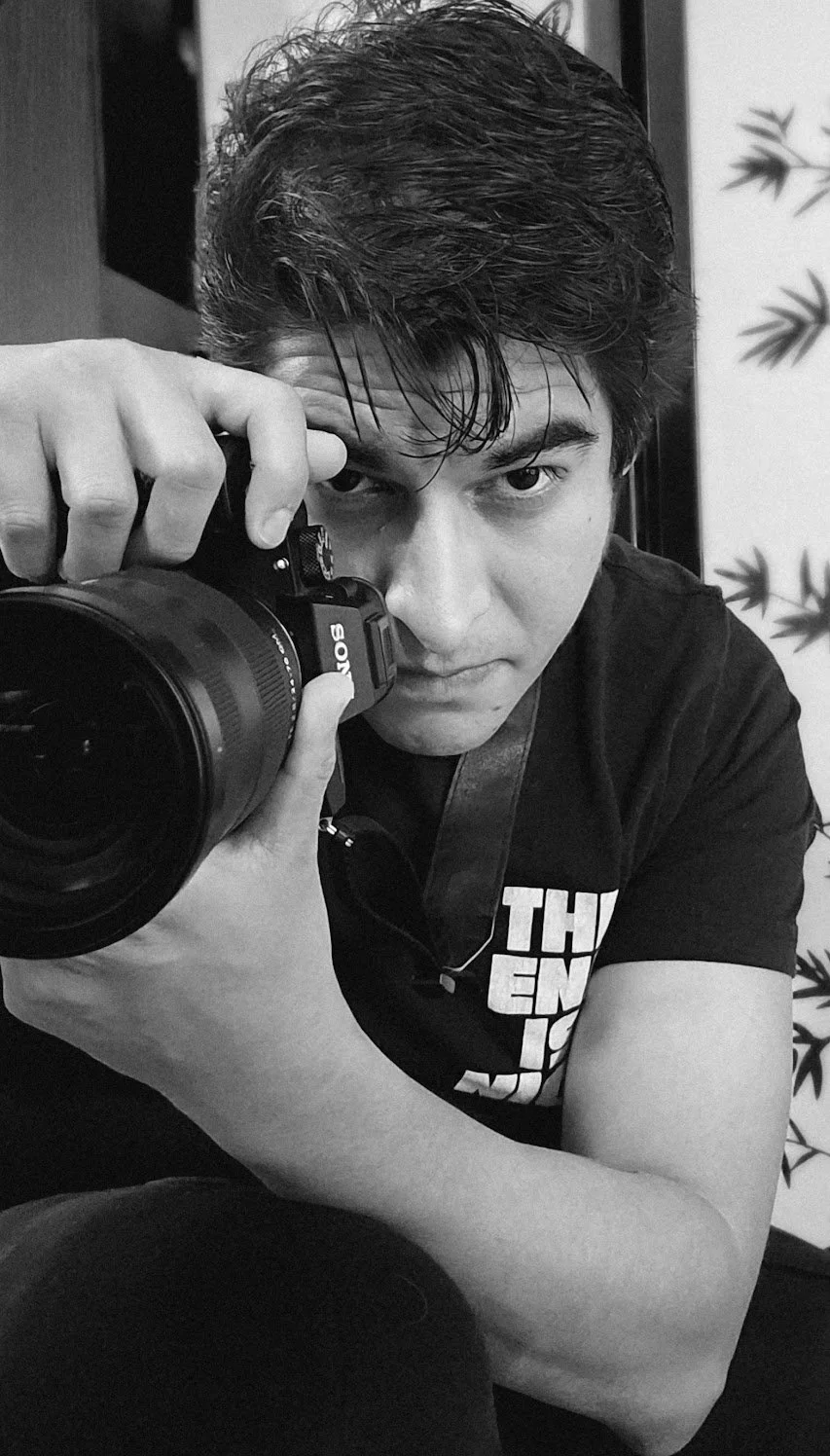 A young man with dark, slightly wet hair taking a selfie with a black Sony camera, looking directly into the lens, in black and white. He is wearing a black t-shirt with white text, and his arm is resting on his knee.