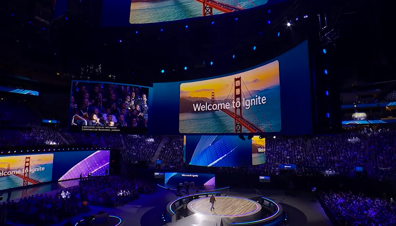 A large conference arena with a stage and big screens displaying a welcome message and images of the Golden Gate Bridge, with an audience seated around the stage.