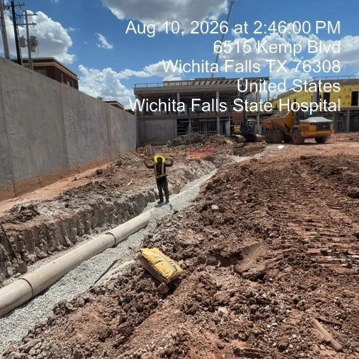 Construction site at Wichita Falls State Hospital with a worker inspecting underground piping, surrounded by dirt and construction equipment under a partly cloudy sky.