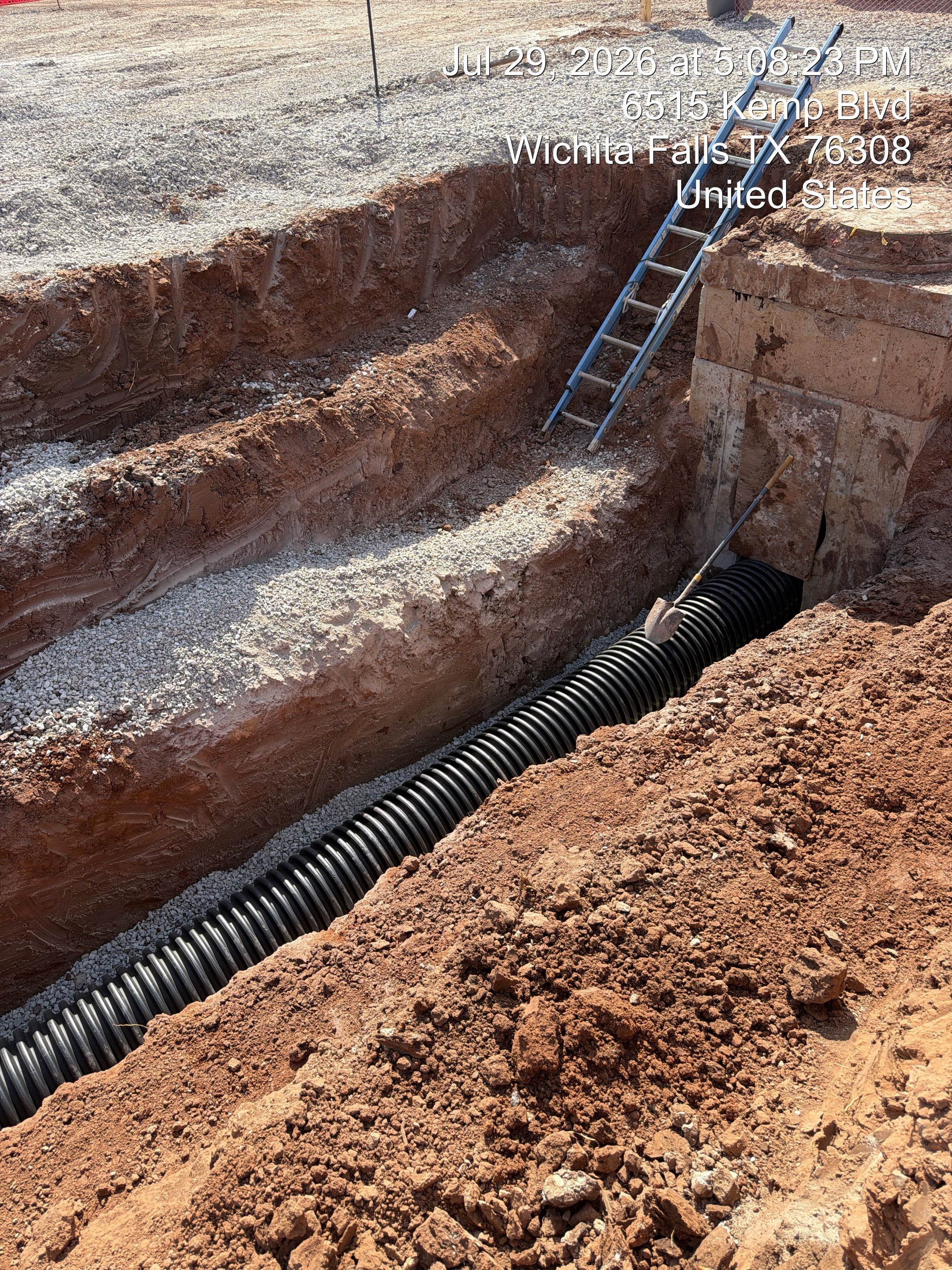 Construction site showing a deep trench with a black corrugated pipe at the bottom, surrounded by reddish-brown dirt and rocks, with a metal ladder leaning against the trench