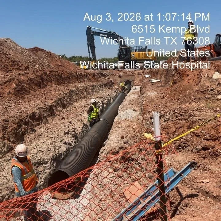 Construction workers installing a large pipe in a deep trench at a work site with an excavator in the background, and orange safety fencing in the foreground.