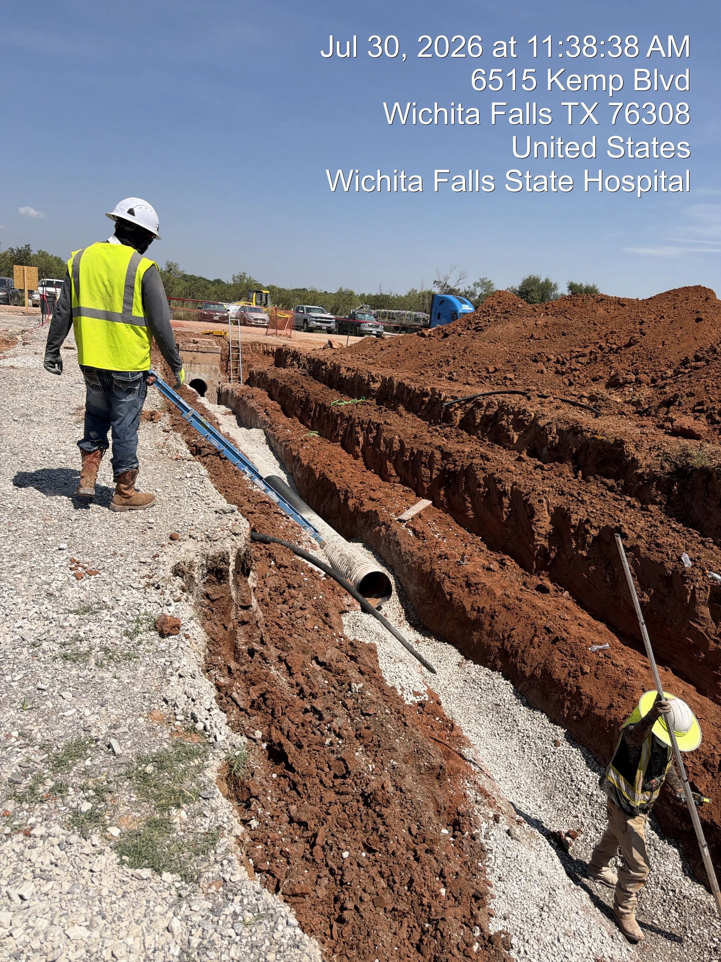 Construction workers at a site with trenches, gravel, a large dirt mound, and parked cars in the background.