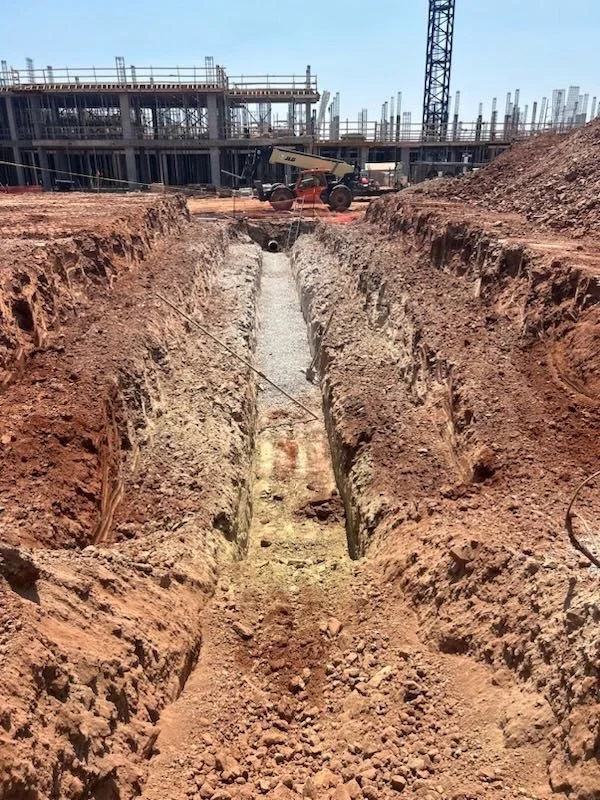 A construction site with an excavation trench running through the dirt, construction equipment on the background, and a crane in the distance.