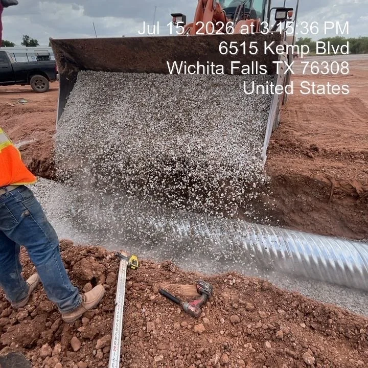 A construction worker is installing a large corrugated metal pipe into a trench, with gravel being dumped into the pipe. The scene is outdoors at a construction site on July 15, 2026, at 3:15 PM, located at 6515 Kemp Blvd, Wichita Falls, TX 76308, United States.