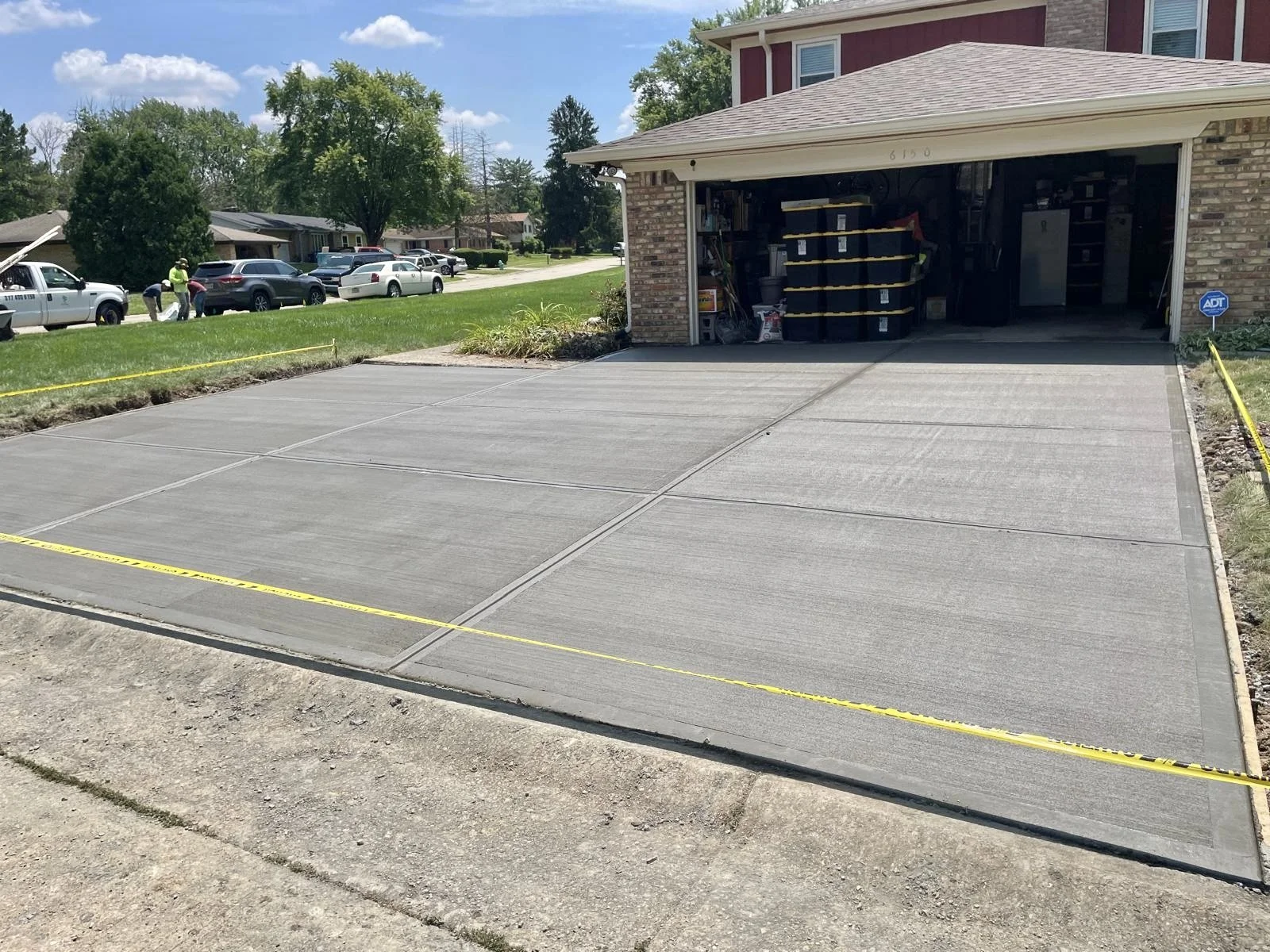 Newly poured concrete driveway in front of a residential garage, with construction caution tape around it. Several cars are parked on the street, and workers can be seen in the background.
