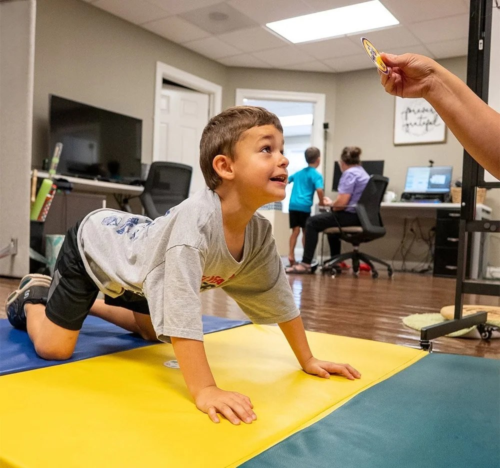 A young boy is crawling on a yellow mat in an office setting, looking up at an adult holding a large badge or token. In the background, two other children and an adult are sitting at desks working on computers.