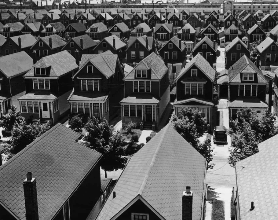 Aerial view of a neighborhood with closely packed houses, mostly two-story homes with gabled roofs, trees lining the streets, and cars parked in driveways.