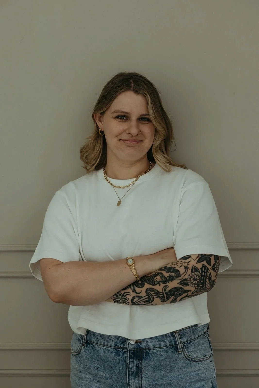 Young woman with blonde hair, tattoos on her left arm, wearing a white t-shirt, denim jeans, and gold jewelry, standing with arms crossed against a beige wall.