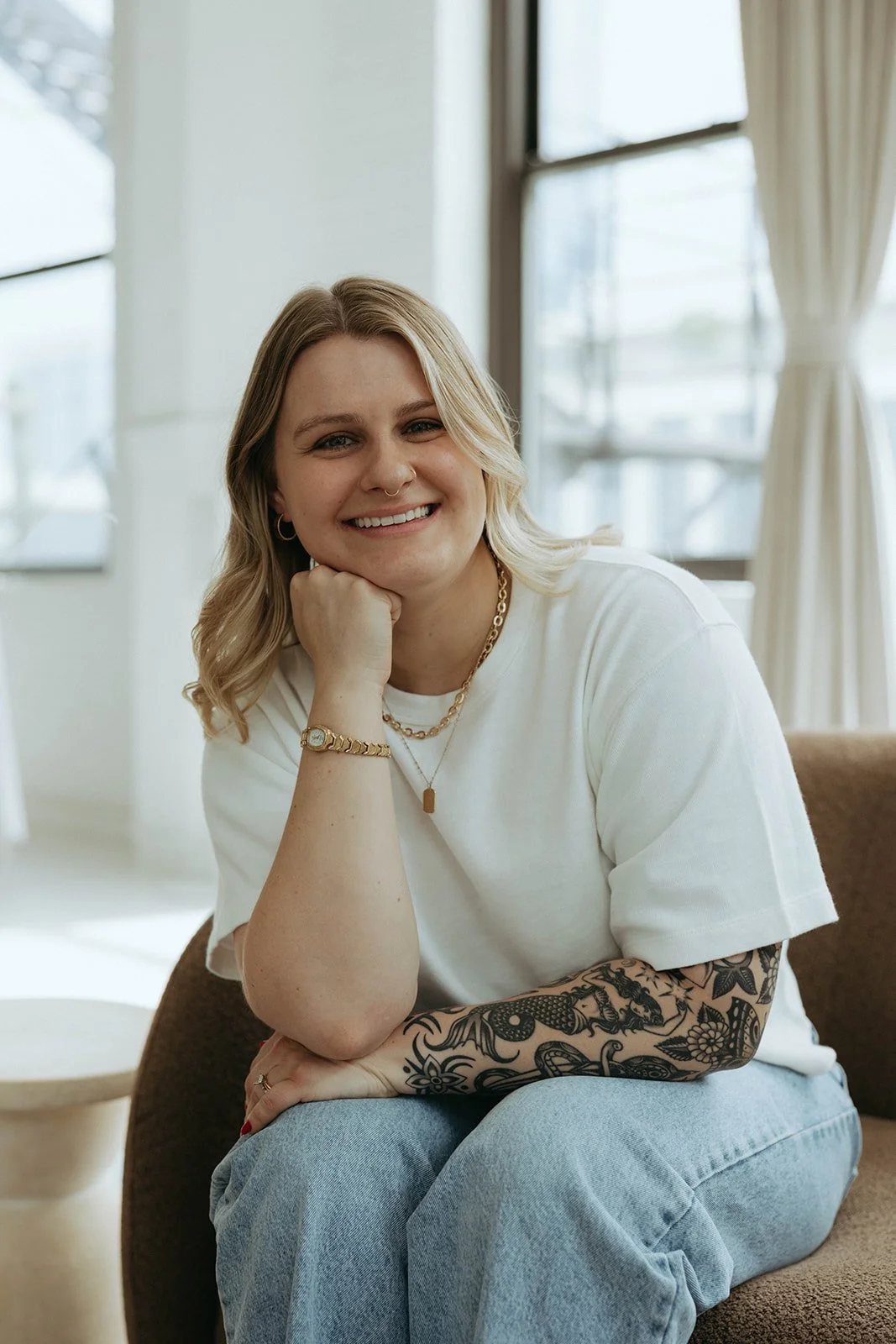 A smiling woman with blonde hair, tattoos on her right arm, wearing a white t-shirt, light blue jeans, jewelry including a necklace, bracelet, and hoop earrings, sitting on a brown chair in a bright room with large windows and cream-colored curtains.