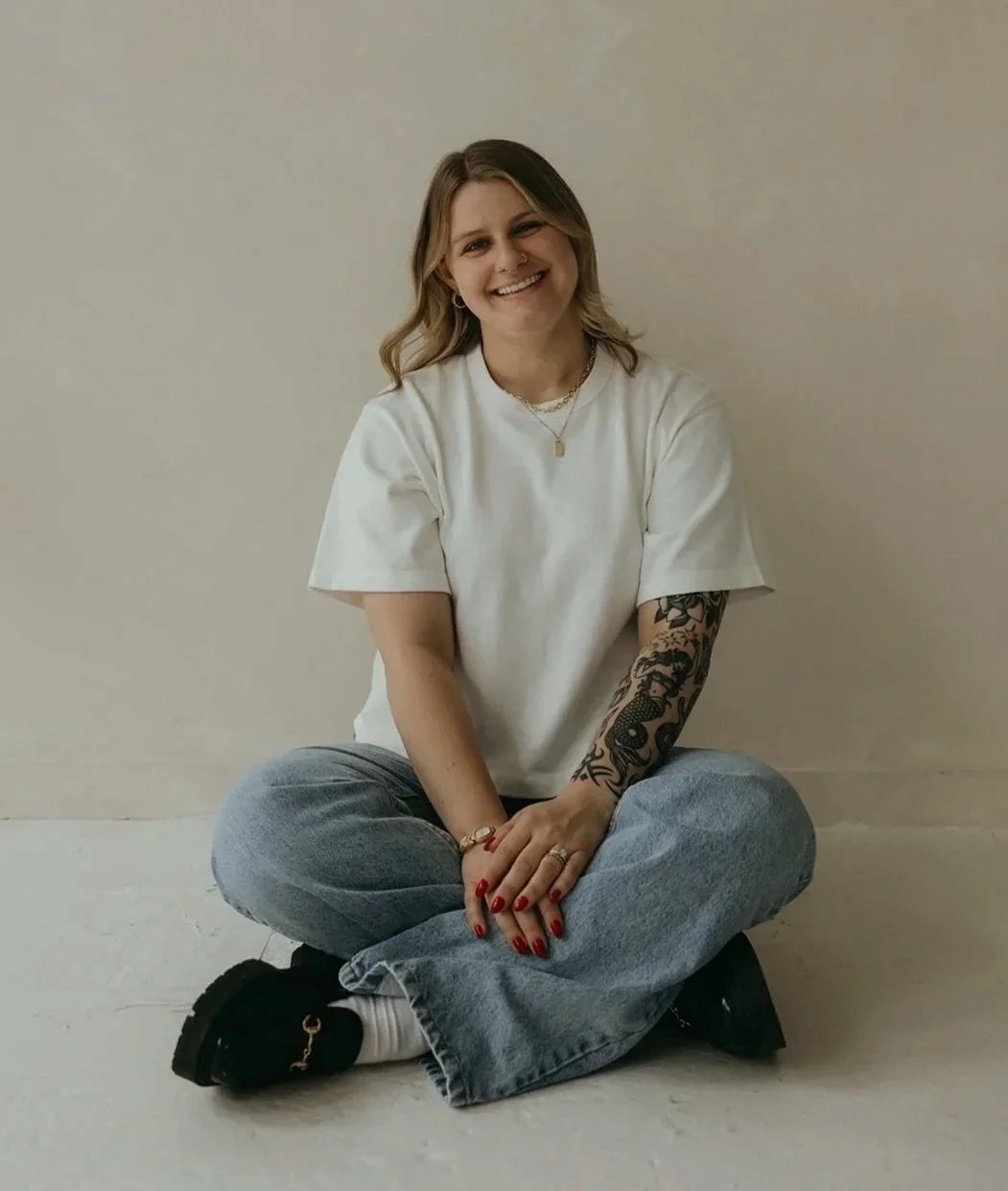 A young woman with blonde hair is sitting cross-legged against a plain beige wall. She is smiling and wearing a white t-shirt, blue jeans, and black shoes. She has a tattoo sleeve on her left arm, and is accessorized with gold jewelry and red nail polish.
