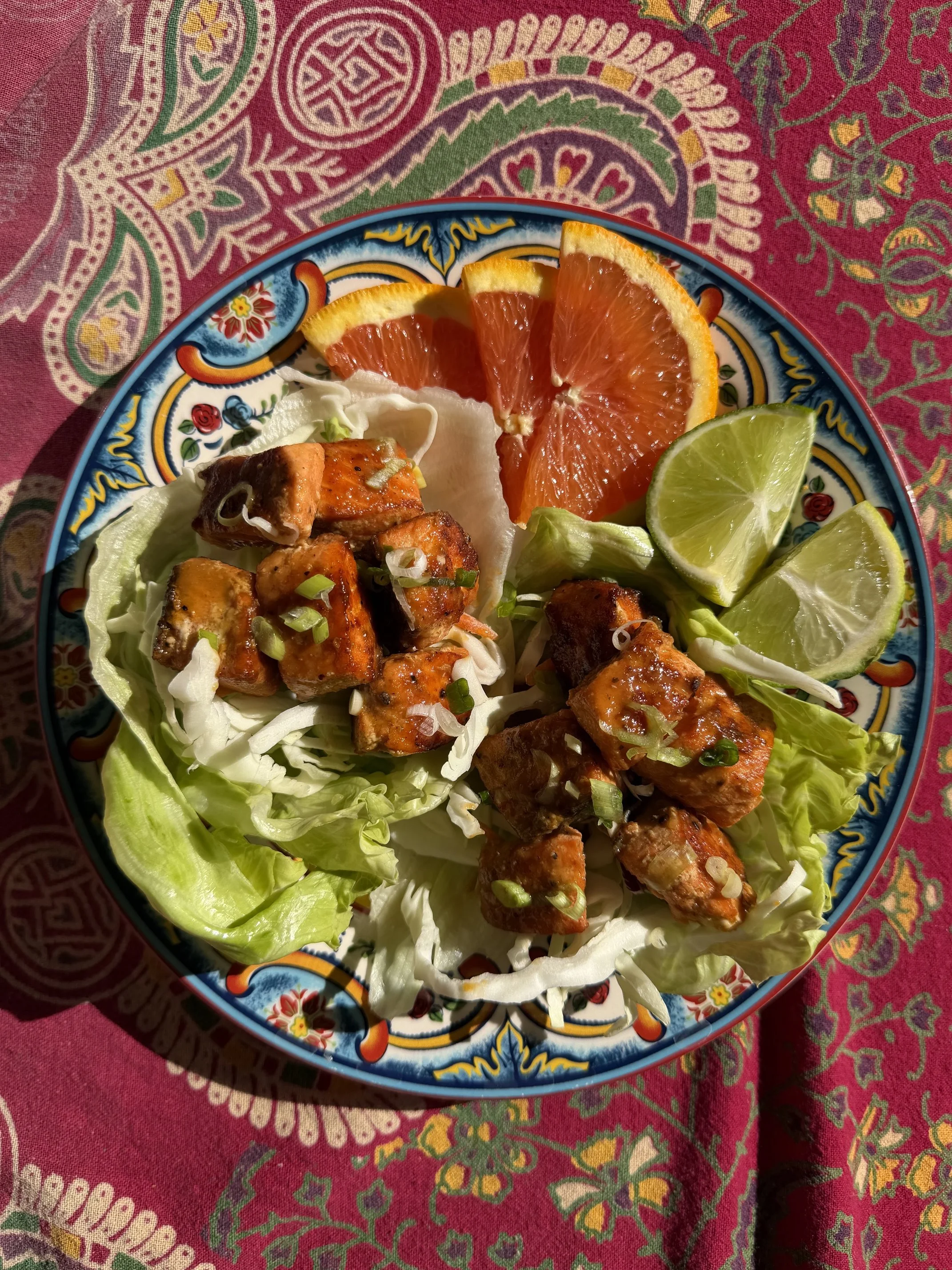 A colorful plate with slices of orange and lime, grilled pieces of salmon, lettuce cups, and some sauce garnished with chopped green onions. The plate is on a red mandala tapestry patterned tablecloth.