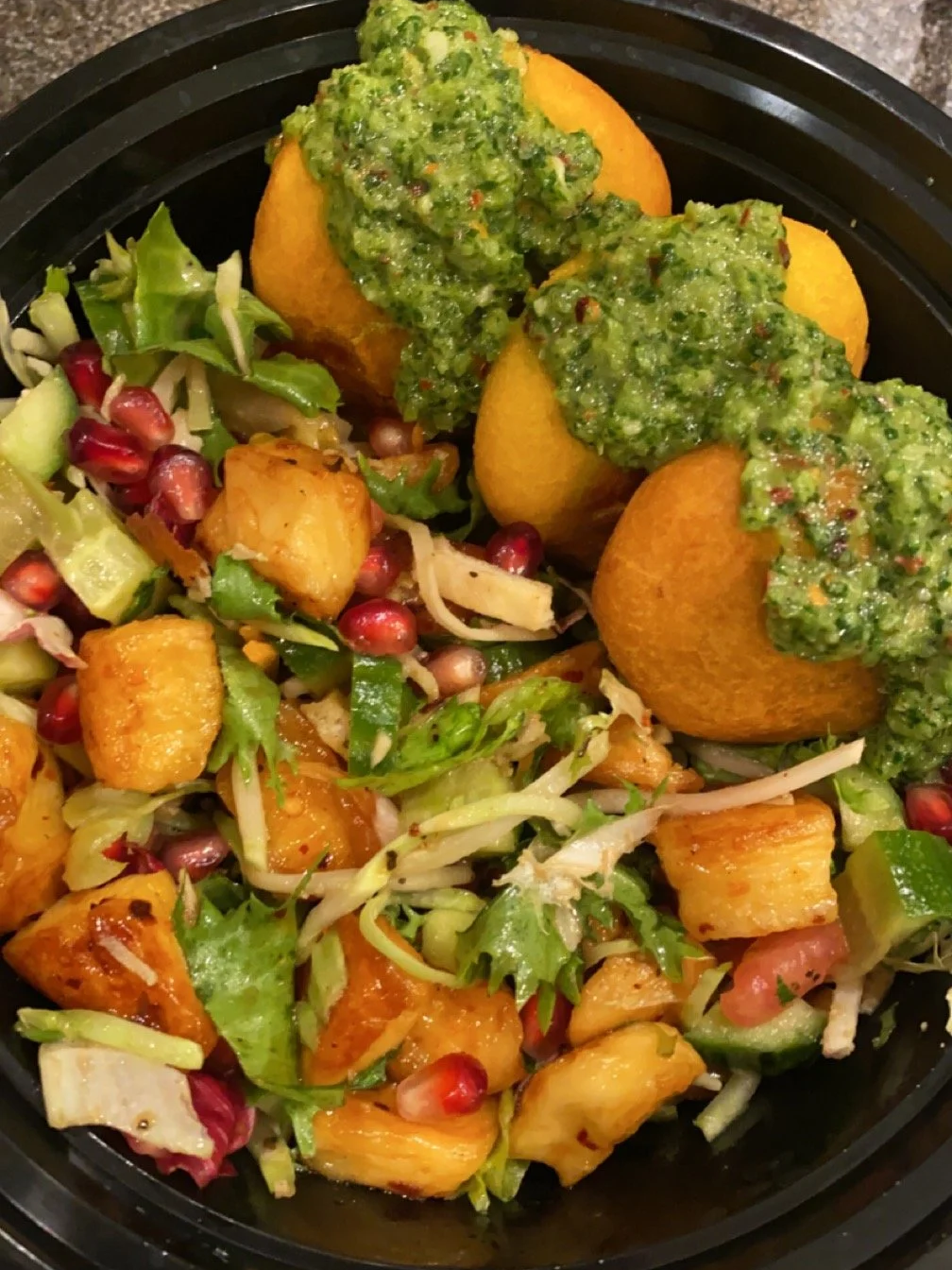 Three fried potato balls topped with green chili sauce, served with a mixed salad including lettuce, pomegranate seeds, cucumber, and chopped vegetables.