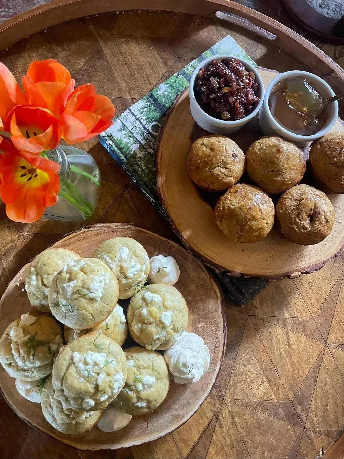 A wooden tray with savory dill muffins topped with homemade whipped honey butter, a smaller wooden tray with carrot muffins and dandelion root jelly, a glass vase with orange tulips, and a magazine with a nature scene.