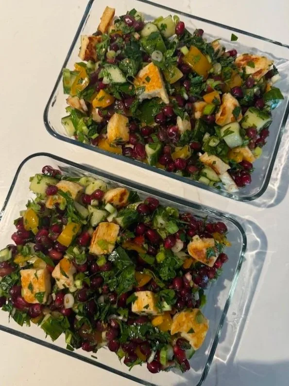 Two glass dishes with chopped salad containing pomegranate seeds, parsley, cucumbers, tomatoes, and grilled hot honey halloumi