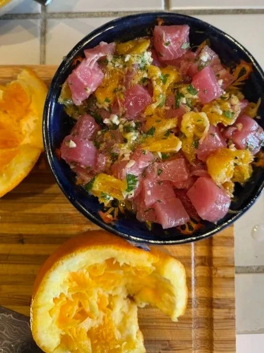 A bowl of tuna poke salad with diced raw tuna, orange, chopped green onions, and sesame seeds, served on a wooden cutting board alongside a slice of orange and a lemon wedge.