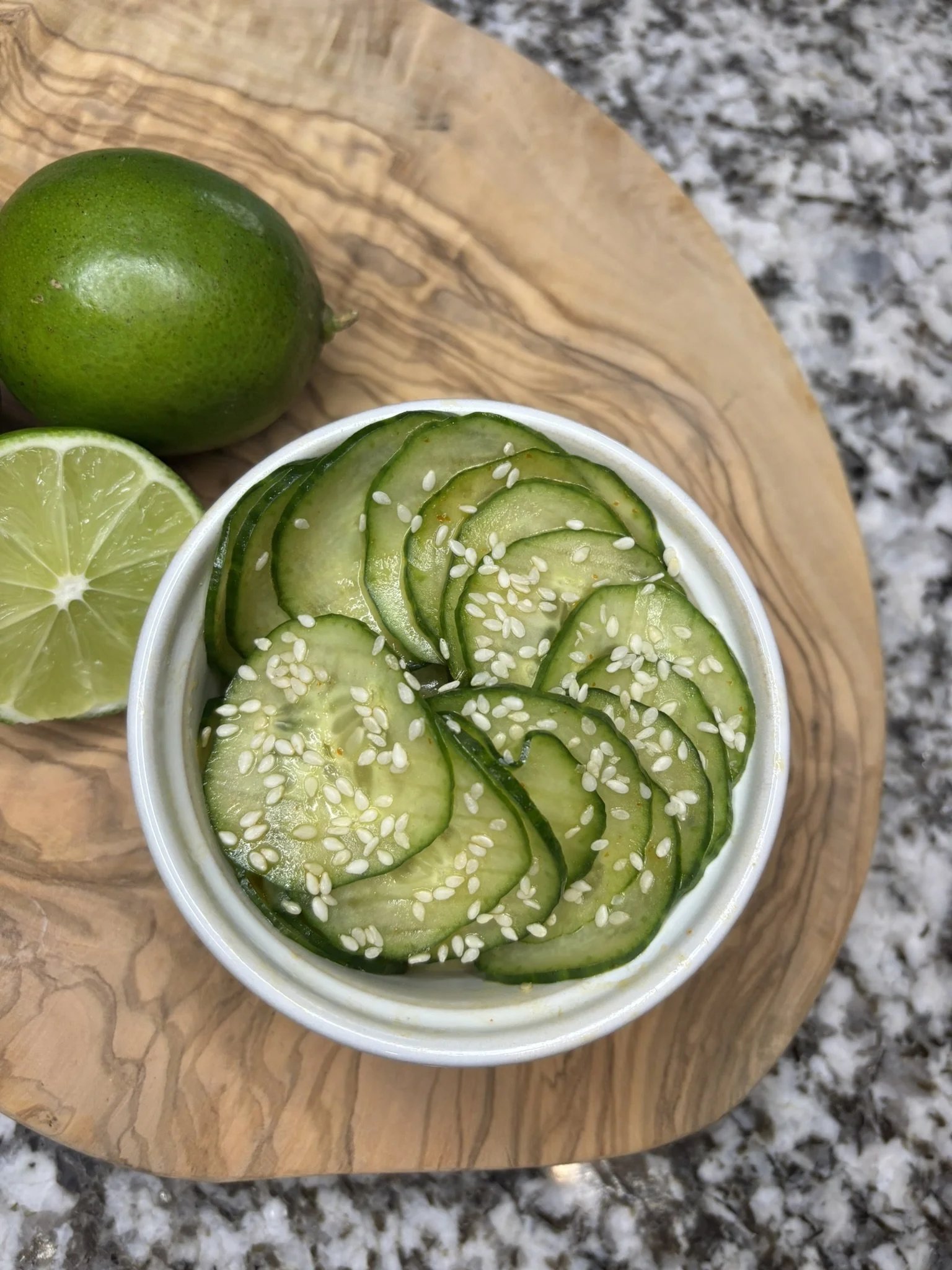 Close-up of a cup of sliced cucumbers sprinkled with sesame seeds, a lime wedge, and a whole lime on a wooden cutting board outside on a granite surface.