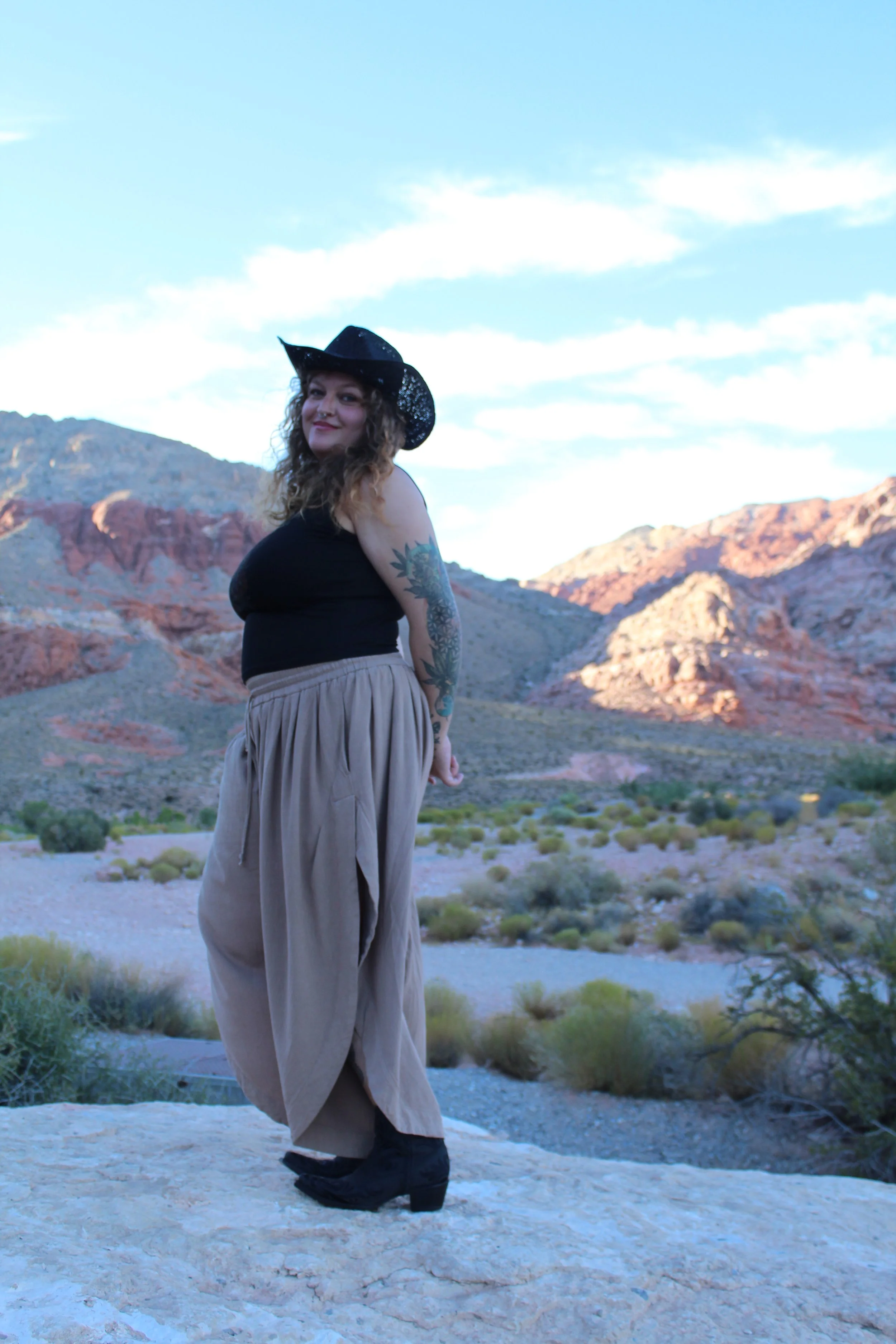 A woman standing on rocky terrain in a desert landscape with colorful rock formations in the background. She is wearing a black cowboy hat, a black top, loose beige pants, and black boots, and is smiling at the camera.