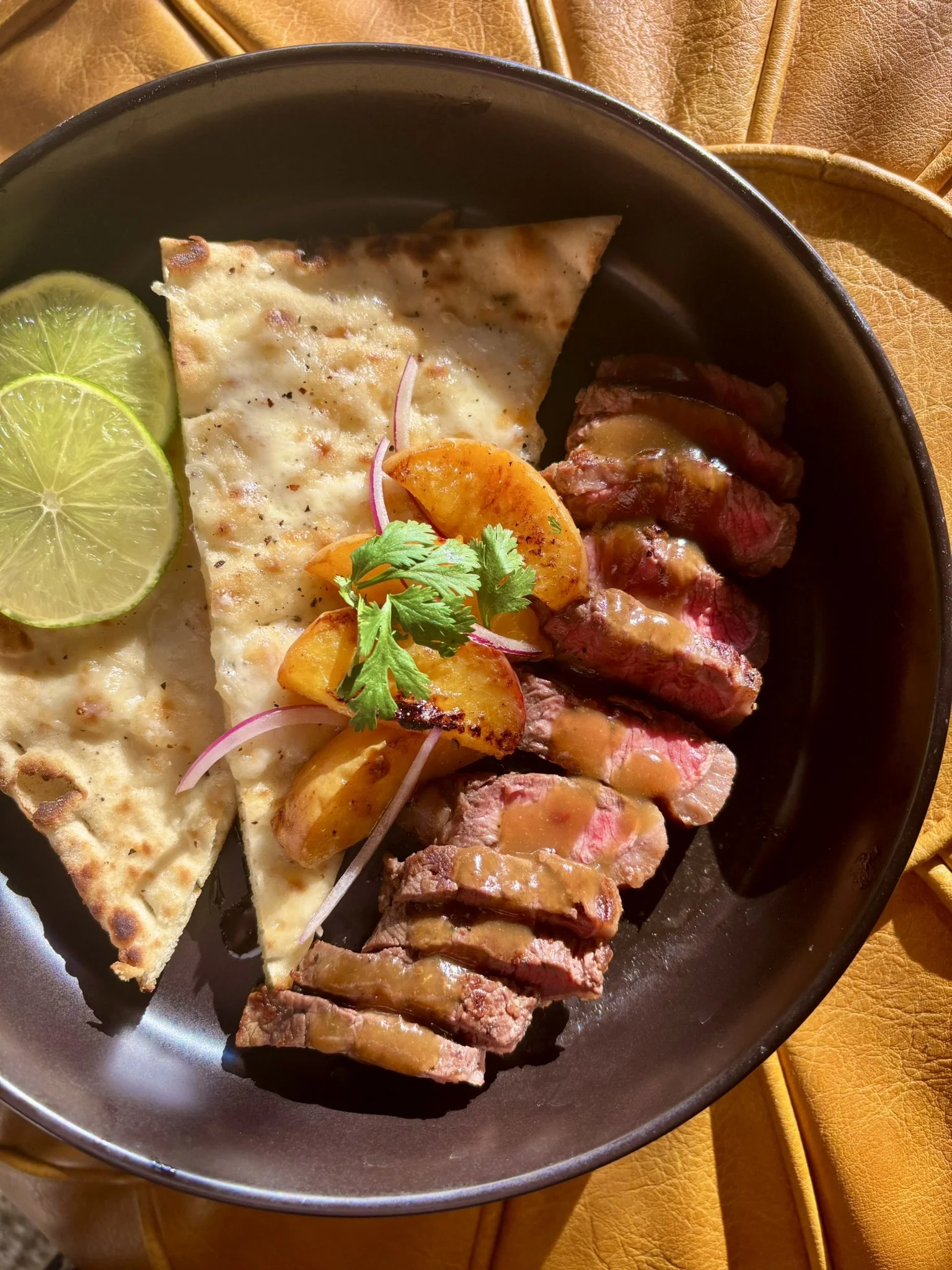 A plate of food containing slices of beef steak with peach glaze, grilled flatbread, lime slices, grilled peaches, and garnished with cilantro, presented on a black plate on a yellow leather surface.