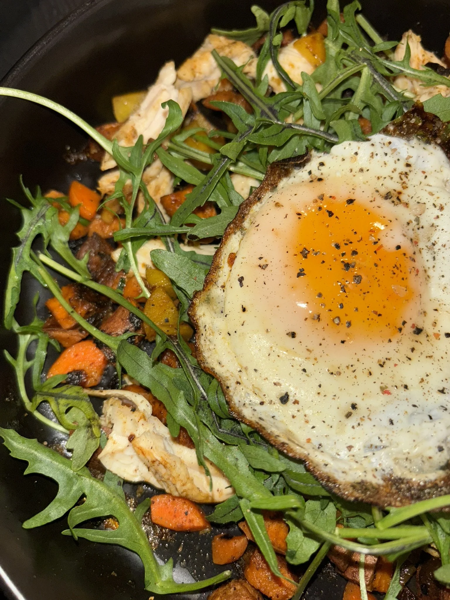 Fried egg cooked sunny side up with black pepper on top, placed on a bed of arugula, grilled vegetables including carrots and sweet potato & onion, and shredded chicken.