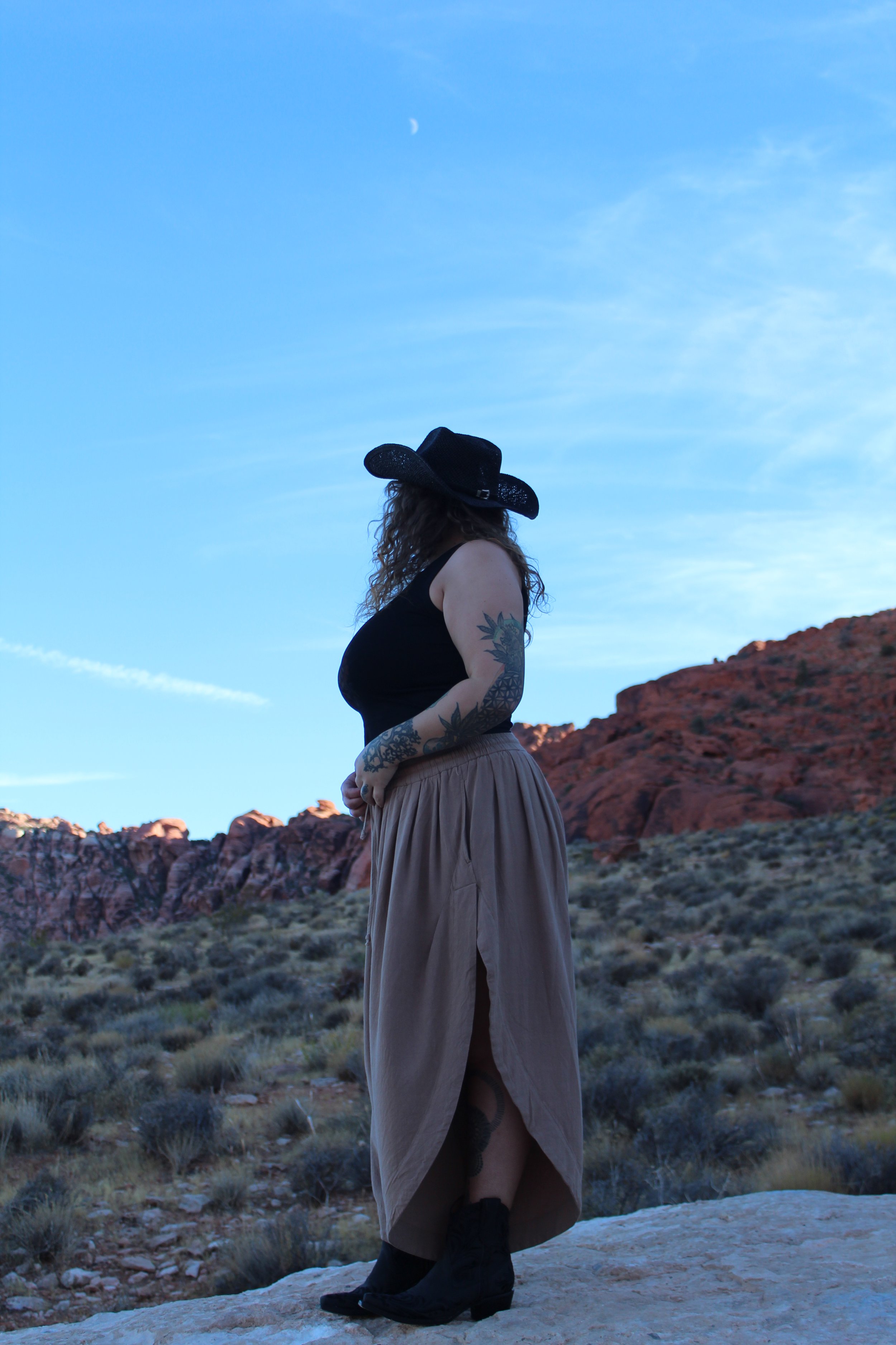 A woman with long curly hair wearing a black wide-brim hat, black tank top, beige skirt, and black boots standing on a rocky terrain with red rock formations in the background under a blue sky with a visible moon.