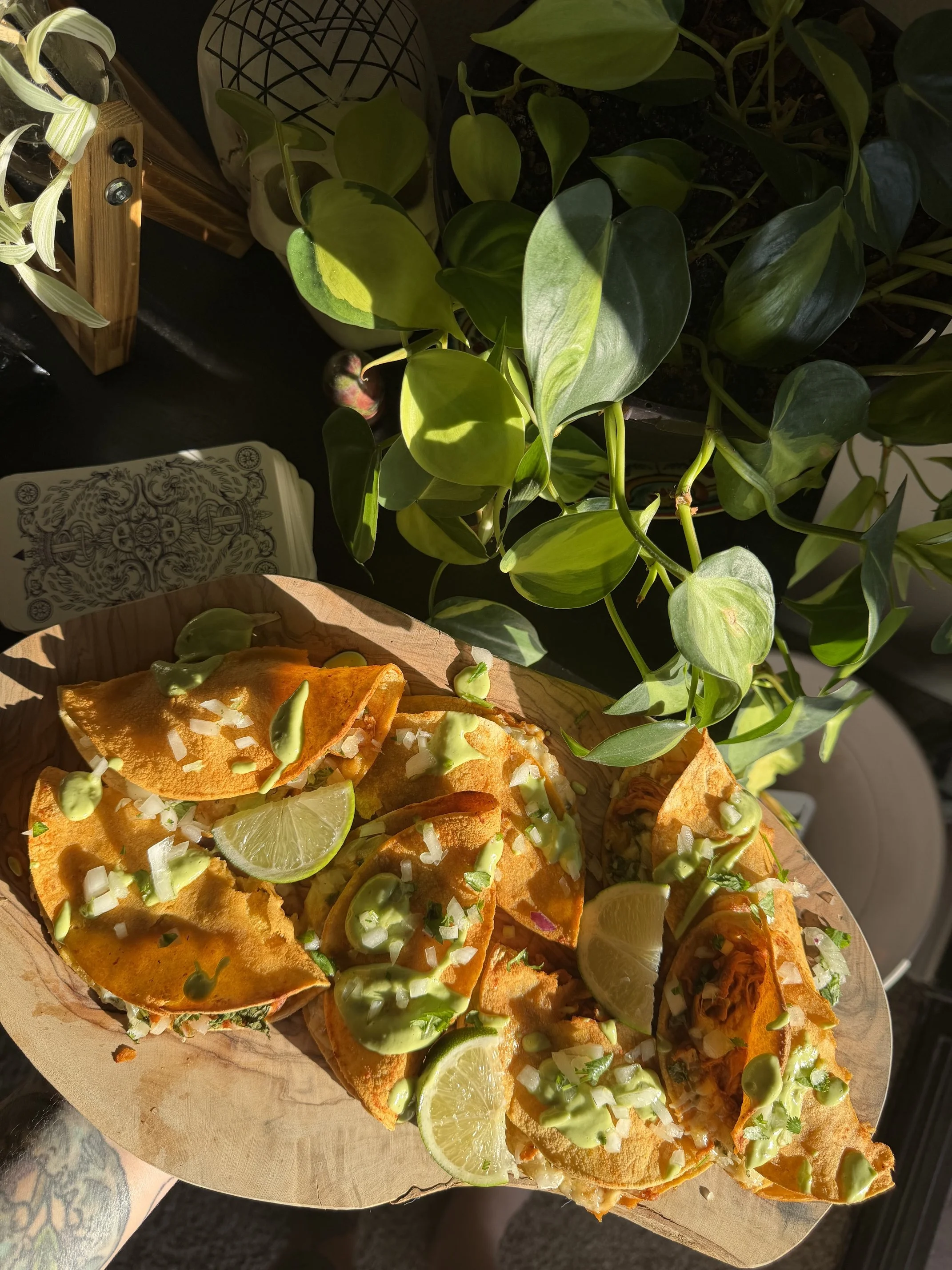 Close-up view of several tacos on a wooden platter, topped with sour cream, chopped onions, sliced lime, and drizzled with green sauce. In the background, there is a potted plant with large green leaves.