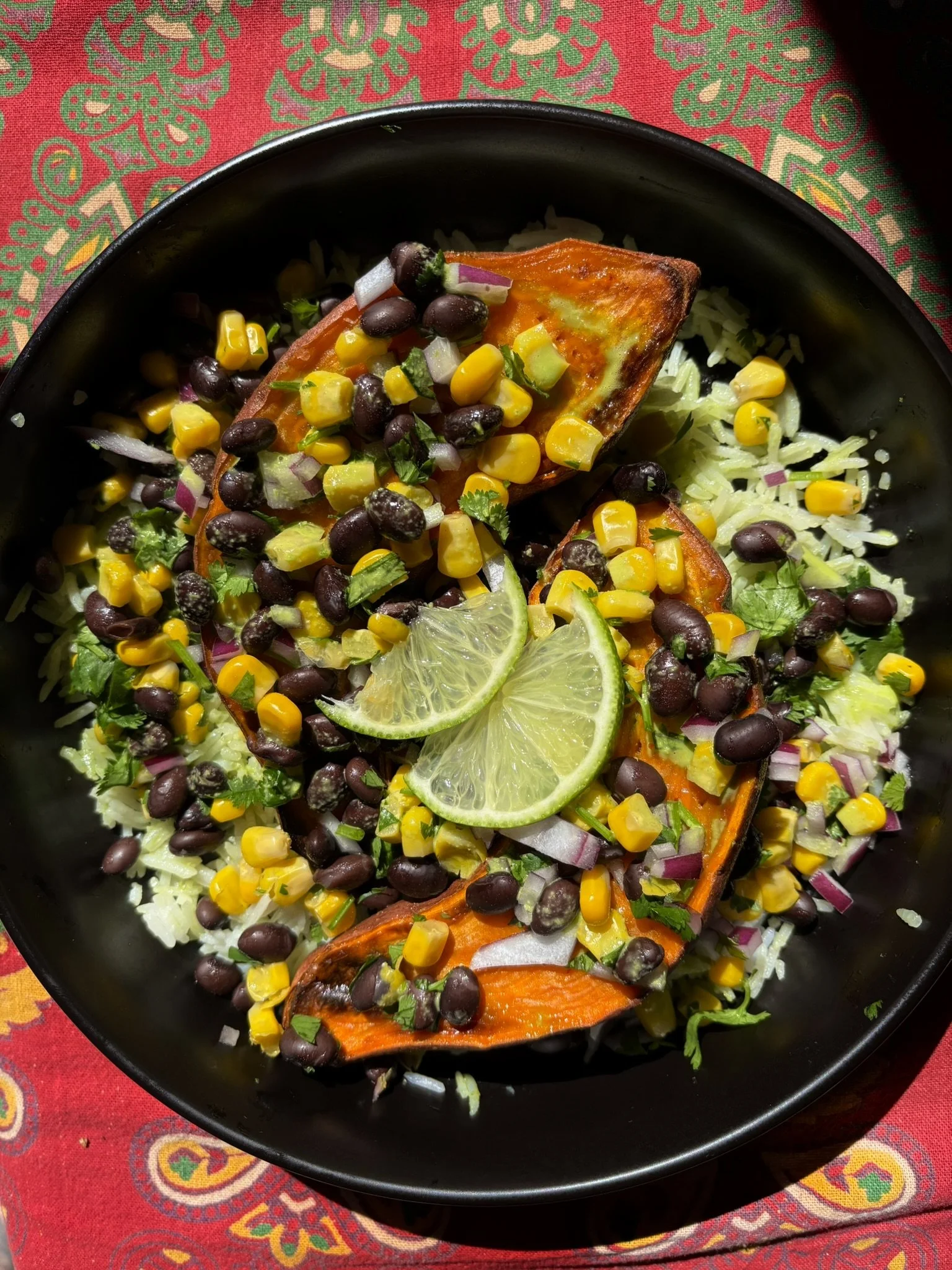 Close-up of a black bowl filled with rice, topped with grilled sweet potato slices, black beans, corn, chopped onions, and fresh lime slices, on a colorful patterned tablecloth.