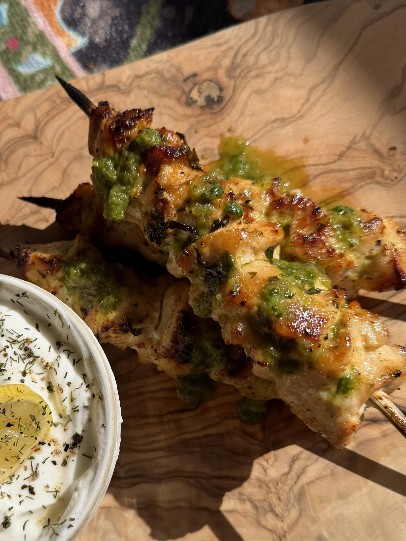 Close-up of a grilled chicken skewer topped with green sauce, served on a wooden platter with a side of creamy sauce garnished with herbs and lemon slices.