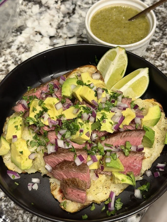 Steak topped with diced avocado, chopped red onion, and chopped cilantro, served with lime wedges, and a side of green salsa over a baked potato