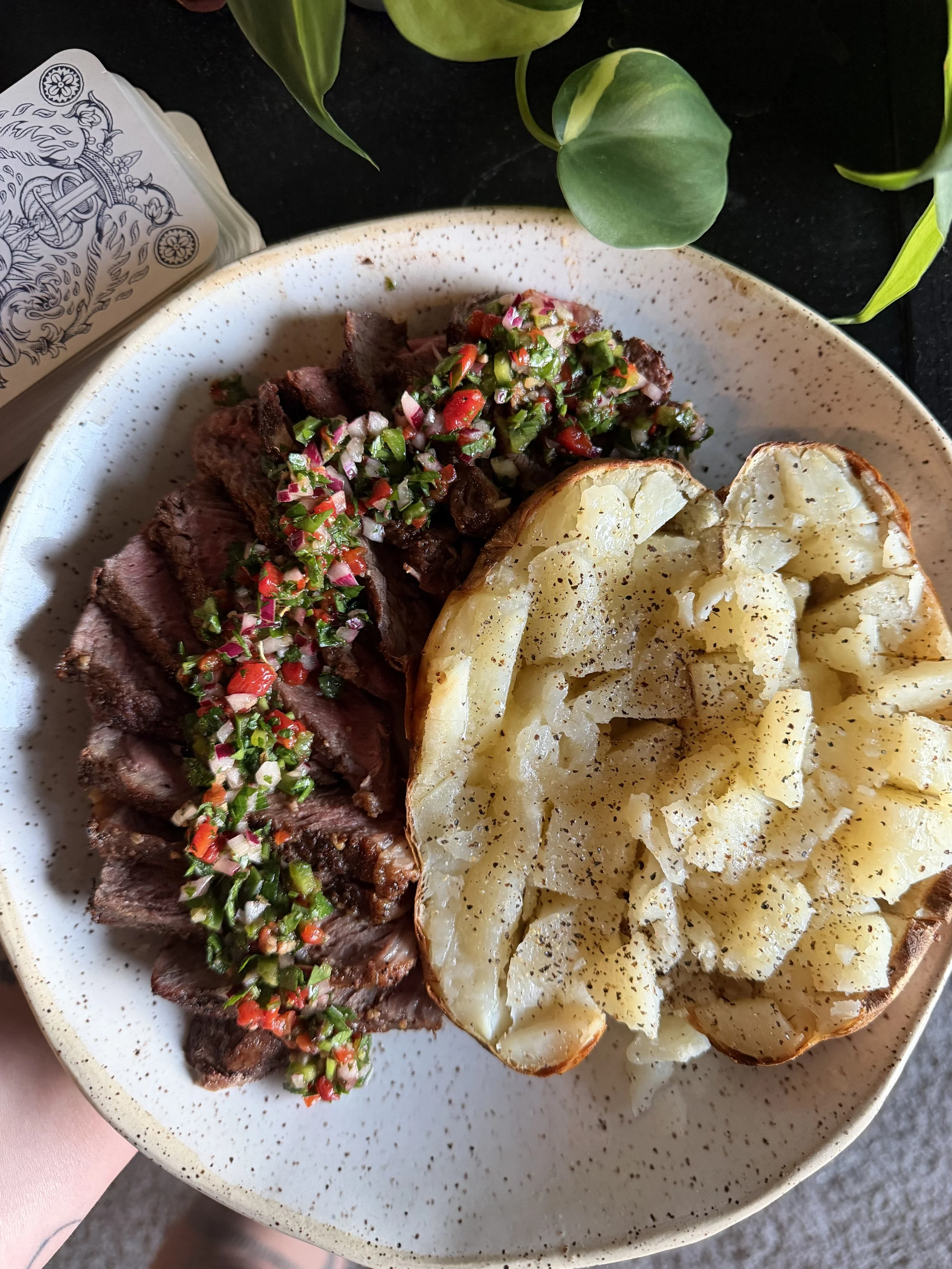 A plate of roasted steak topped with a fresh chimichurri of chopped red chili, onion, and herbs, accompanied by sliced baked potato halves sprinkled with black pepper.