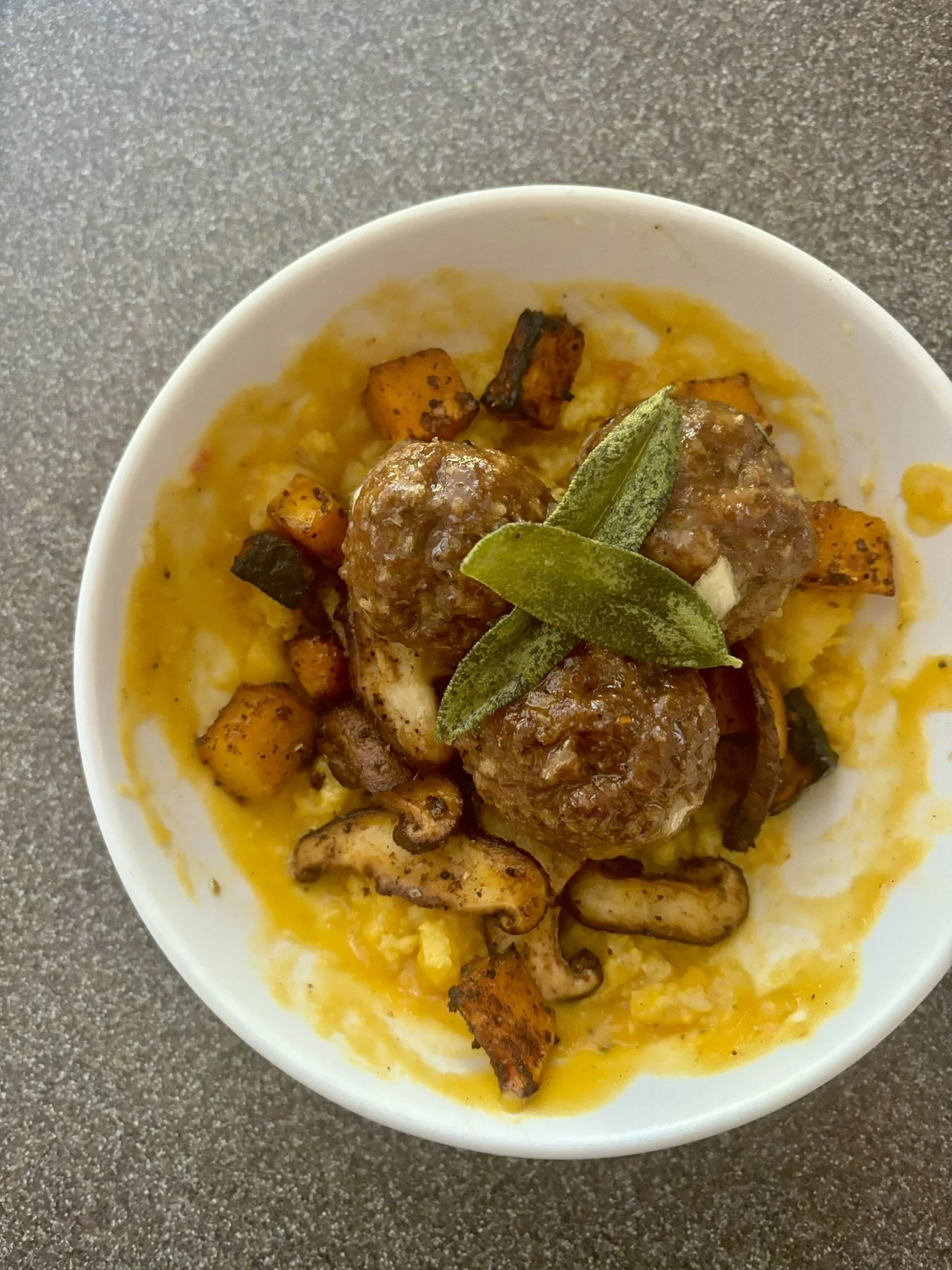 Close-up of a bowl of sage meatballs, butternut squash sauce, fried sage, served on a textured gray surface.