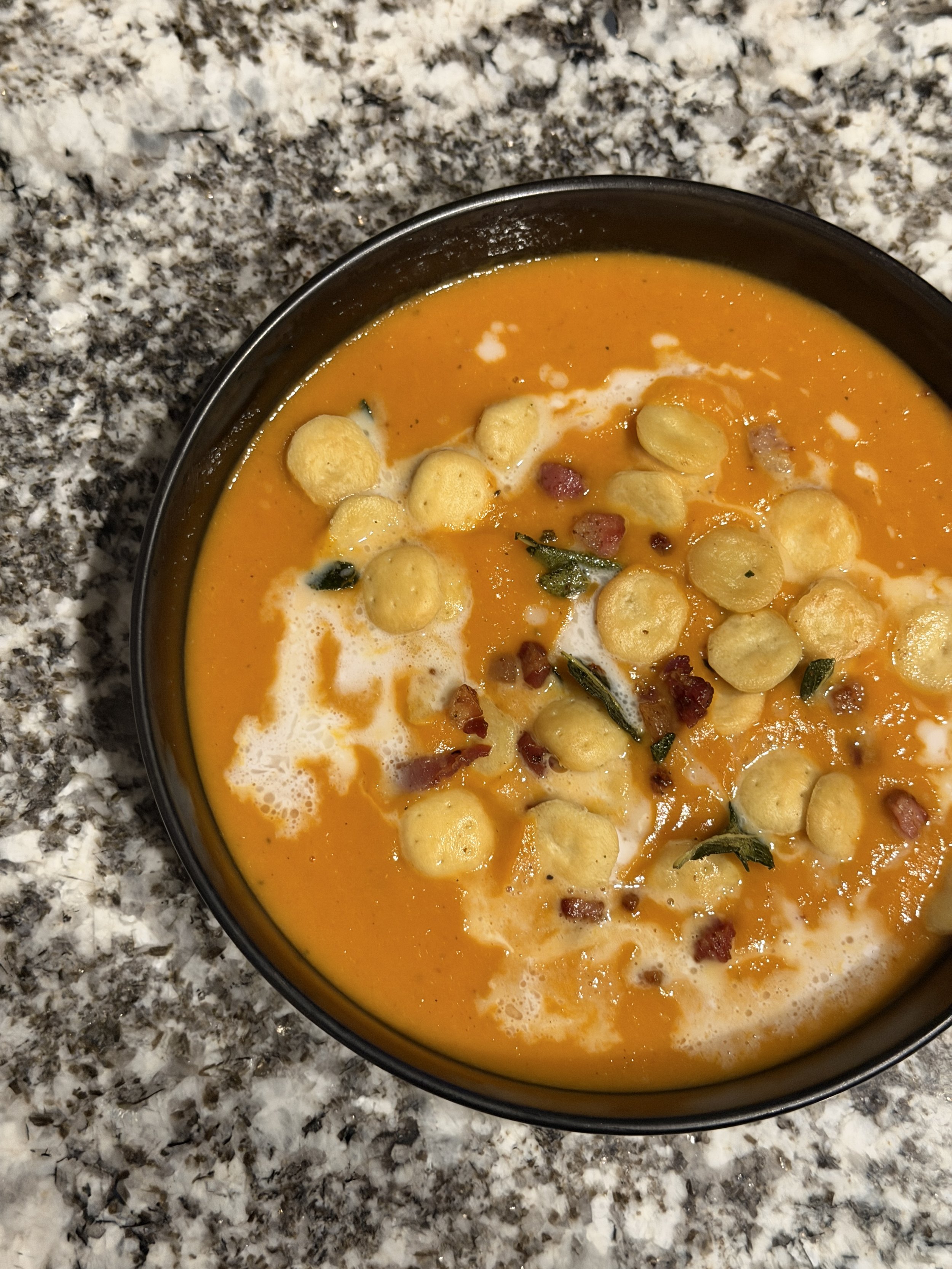 A bowl of creamy butternut squash soup topped with oyster crackers, pancetta, and fried sage, drizzled with coconut milk placed on a granite countertop.
