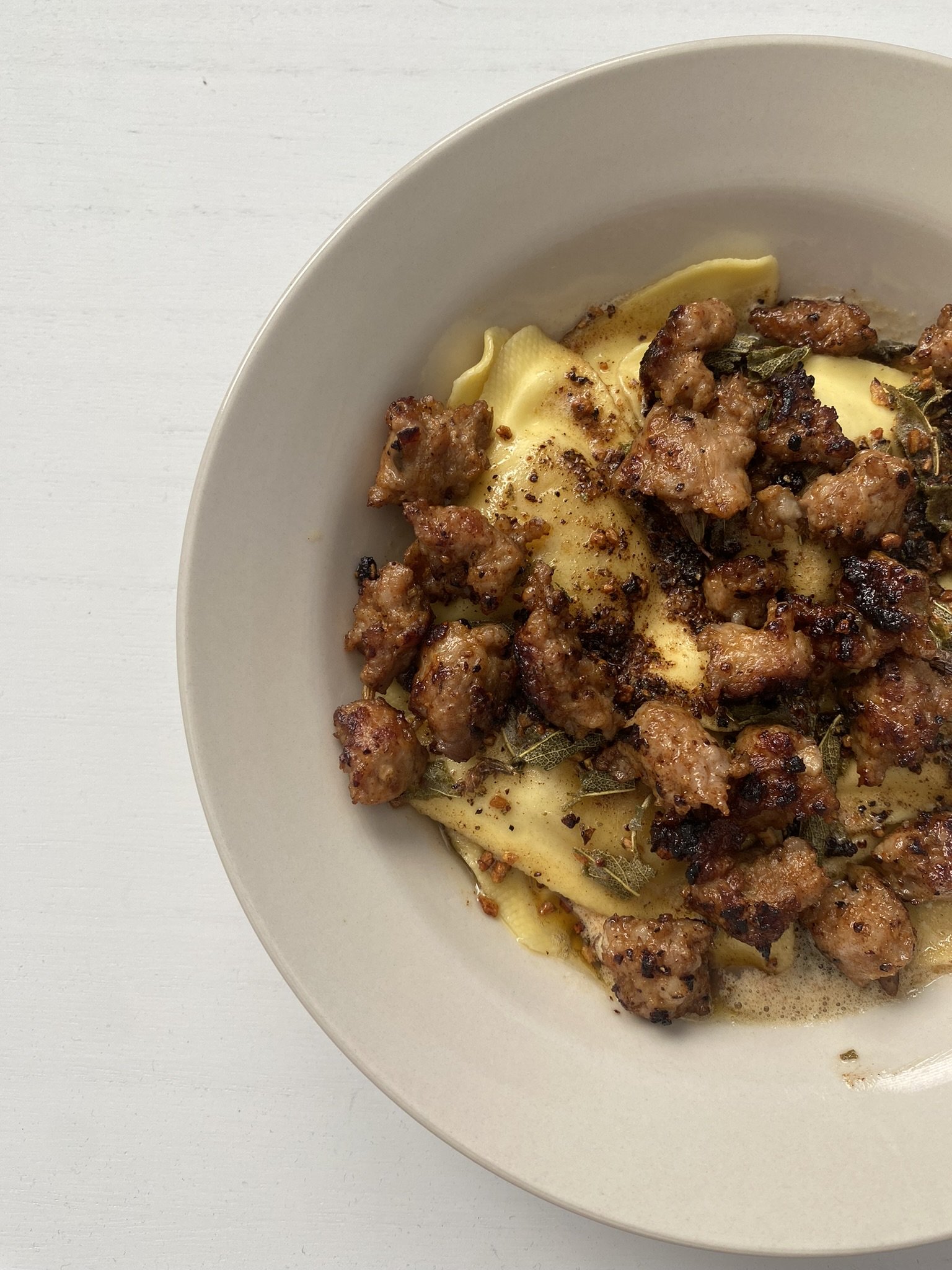 A white bowl contains cooked ravioli pasta topped with sweet italian sausage and herbs, garnished with spices on a white surface.