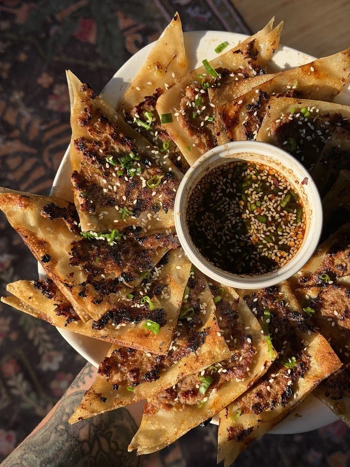 A plate of fried dumplings topped with green onions and sesame seeds, served with a small cup of soy sauce dipping sauce.