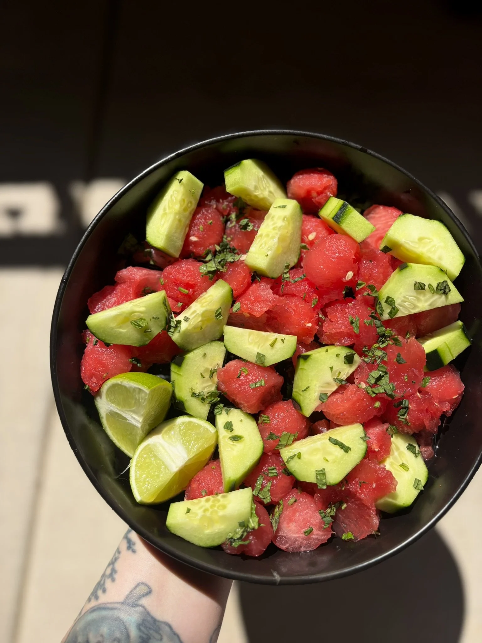 A black bowl filled with chopped cucumbers, watermelon, lime wedges, and garnished with herbs, held by a person with a tattooed arm.
