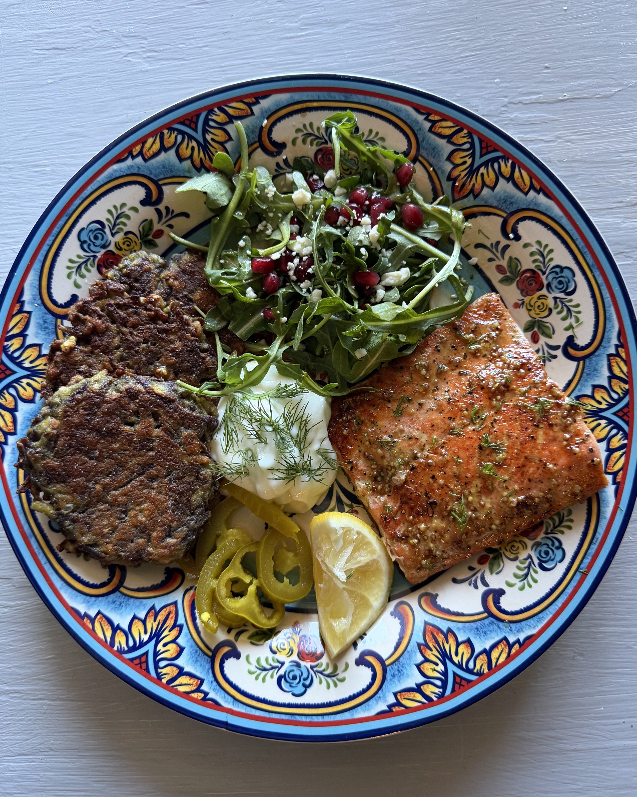 Plate with grilled purple carrot fritters, salmon, and side salad with pomegranate seeds, arugula, and crumbled cheese, lemon dill yogurt sauce, garnished with lemon slices.