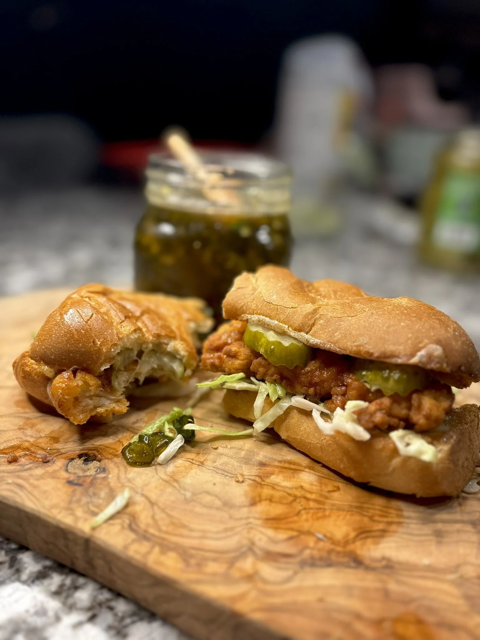Close-up of a fried chicken sandwich with lettuce and candied jalapenos on a wooden serving board, with a jar of jalapeno honey sauce and other condiments in the background.